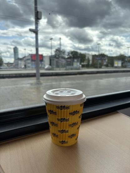 Ein Einwegbecher auf einem Tisch im einem Zug, im Hintergrund sind Bahnsteige und Gebäude zu sehen