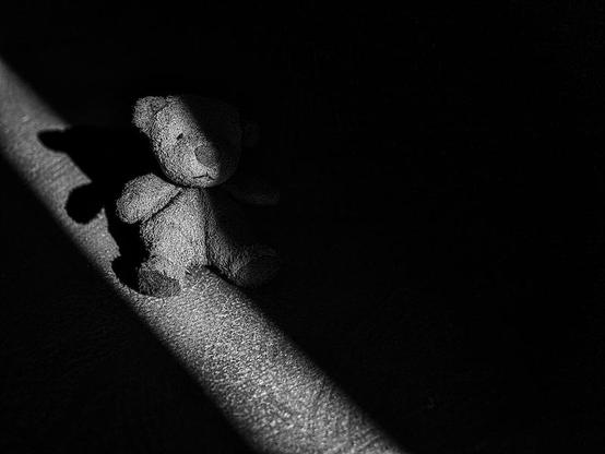 Man sieht einen kleinen Teddy, der durch das Licht von einem Türspalt ca. zur Hälfte angestrahlt wird. Das Foto ist in Schwarzweiß.