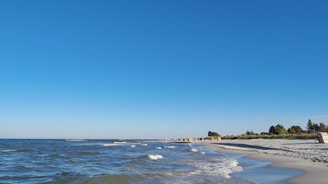 Foto, quer. Blick über Meer und Küste.
Von links bewegt sich das Meer mit Wellen und Gischtauf den Strand zu, der rechts in einem sanften Linksbogen gen Horizont läuft. Vereinzelt ein paar Strandkörbe, rechts mittig ein paar Bäume, über allen ein wolkenloser blauer Himmel.