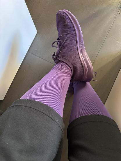 Übereinandergeschlagene Beine auf einem dunklen Boden. Sowohl Schuhe als auch Strümpfe in violett sind sichtbar, darüber eine schwarze Hose