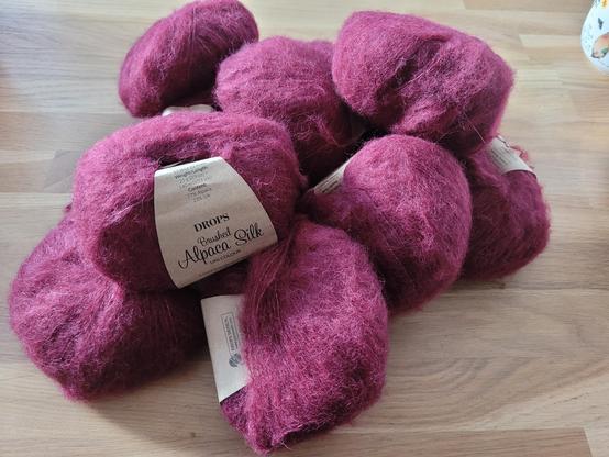 Ein Haufen sehr flauschiger Wollknäuele liegt auf einer Holzplatte. Die Wollknäuele sind weinrot. Von einem Knäuel  ist das Etikett sichtbar. Darauf steht "Drops - Brushed Alpaca Silk"