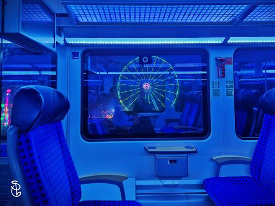 Das Photo zeigt ein S-Bahn-Fenster. Im Fenster ist ein beleuchtetes Riesenrad zu sehen. Die Lichtstimmung ist blau.