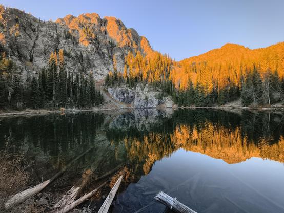 Sunrise alpenglow on rocky peaks beyond a reflective lake bordered by pine trees.