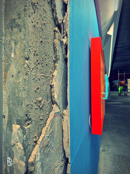 Unter dem Putz einer Wand sind offene Stellen und loser Mörtel. Die Wand - links im Bild - ist blau, auf ihr ein Bilderrahmen in orange. Nach rechts öffnet sich der Blick in den Raum.