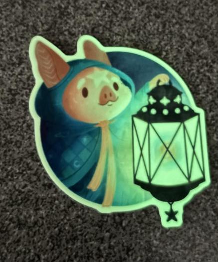Ein Comic-Schweinchen mit Umhang und Laterne. Der Aufkleber ist im Dunklen und leuchtet grün.