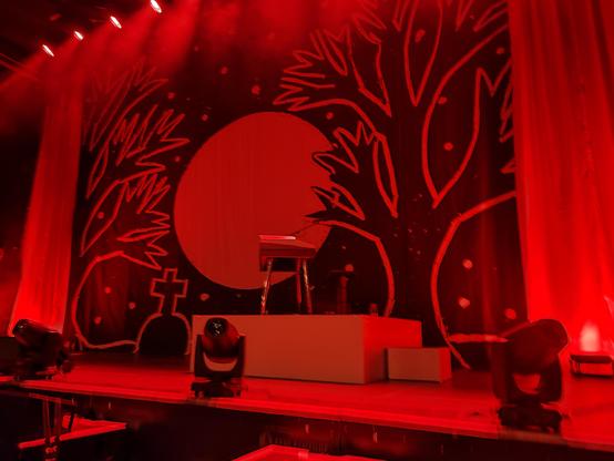 In rotes Licht getauchte Bühne

Linoleum Druck als Hintergrund Banner

Ein Baum an jeder Seite
Mond sterne in der Mitte
Grab Links 

In schwarz weiß 

Podest mit e-Piano und Stuhl