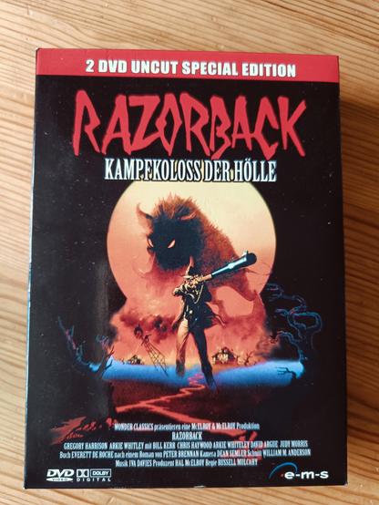 DVD-Cover des Films "Razorback". Deutscher Untertitel: "Kampfkoloss der Hölle". Eine Typ mit einem riesigen Gewehr ist zu sehen, im Hintergrund ein noch riesigerest Wildschwein.