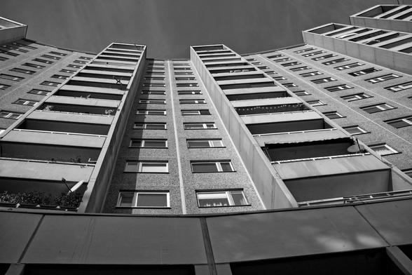 Schwarzweiß Foto, Blick nach oben an einem Hochhaus. Fenster und Balkone von unten. Ganz unten die Betonteile der Eingänge.