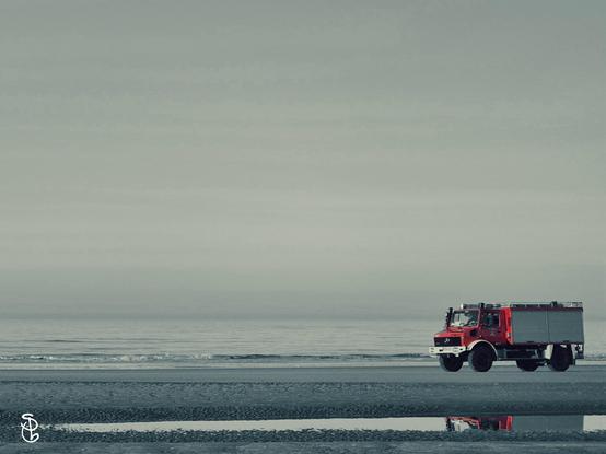 Ein Feuerwehrauto fährt am Strand der Nordsee entlang. Im Hintergrund sieht man das Meer und den Himmel. Im Vordergrund spiegelt sich ein Teil des LKW in einer Pfütze, die von der Ebbe zurückgelassen wurde.