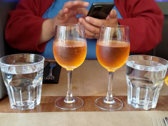 Zwei Gläser Cidre in der Mitte, rechts und links daneben je ein Glas Wasser. Im Hintergrund sitzt Tom und tippt auf seinem Handy. 