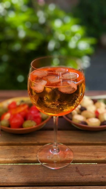 Ein Glas Aperol Spritz, dahinter zwei Schalen mit Antipasti wie Oliven, gefüllte Kirschpaprika.