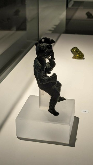Kleine Bronzestatue von der Seite, die Figur sitzt, auf Stirn und als Nase ist jeweils deutlich ein Phallus zu sehen.