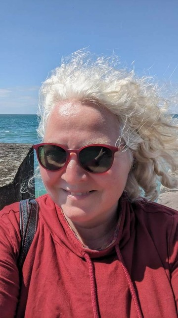 Ich lächele in die Kamera und habe eine Sonnenbrille auf. Meine Haare wehen im Wind, im Hintergrund ist Meer zu sehen.