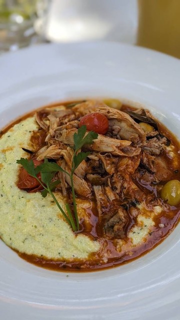 Tiefer weißer Teller, darin auf einer Seite gelbe Polenta, rechts daneben Kaninchenfleisch in rötlicher Sauce, kleine Tomaten, grüne Oliven. 