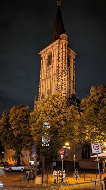 Gotischer Kirchturm an der Fassade der Kirche, viele Bäume stehen auf dem Platz vor der Kirche, die in der Dunkelheit beleuchtet ist. 