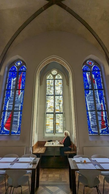 Eine Altarnische, links und rechts bemalte neogotische hohe Fenster, darüber sieht man noch die Rippen eines Gewölbes. In der Nische ein Tisch, daran sitzt eine Frau und frühstückt. 